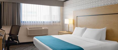 A hotel room with a large bed, a desk with a chair, a lamp, and a window with curtains.