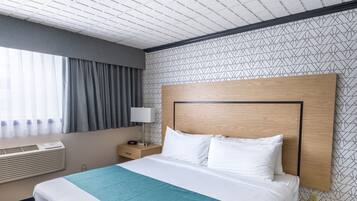 A hotel room with a large bed, a wooden headboard, a nightstand with a lamp, a window with curtains, and a ceiling with a grid pattern.