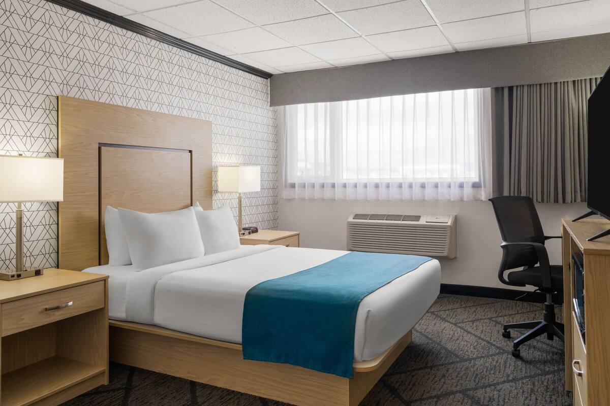 A hotel room with a bed, two bedside tables with lamps, a desk with a chair, and a window with curtains.