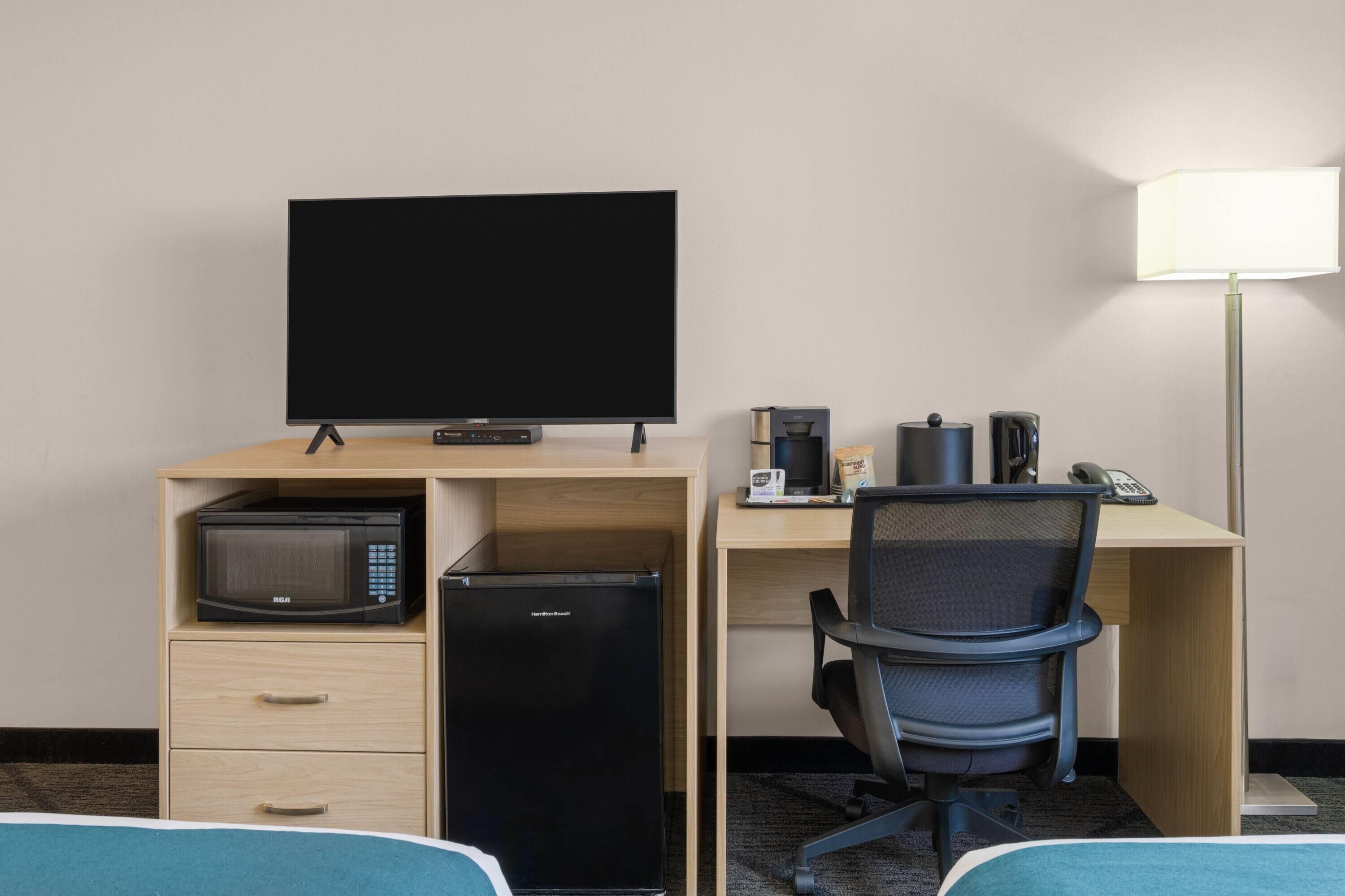 Habitación de hotel con cama, escritorio con computadora, microondas, refrigerador, televisor, lámpara y cafetera.