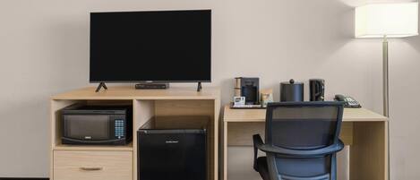Habitación de hotel con cama, escritorio con computadora, microondas, refrigerador, televisor, lámpara y cafetera.