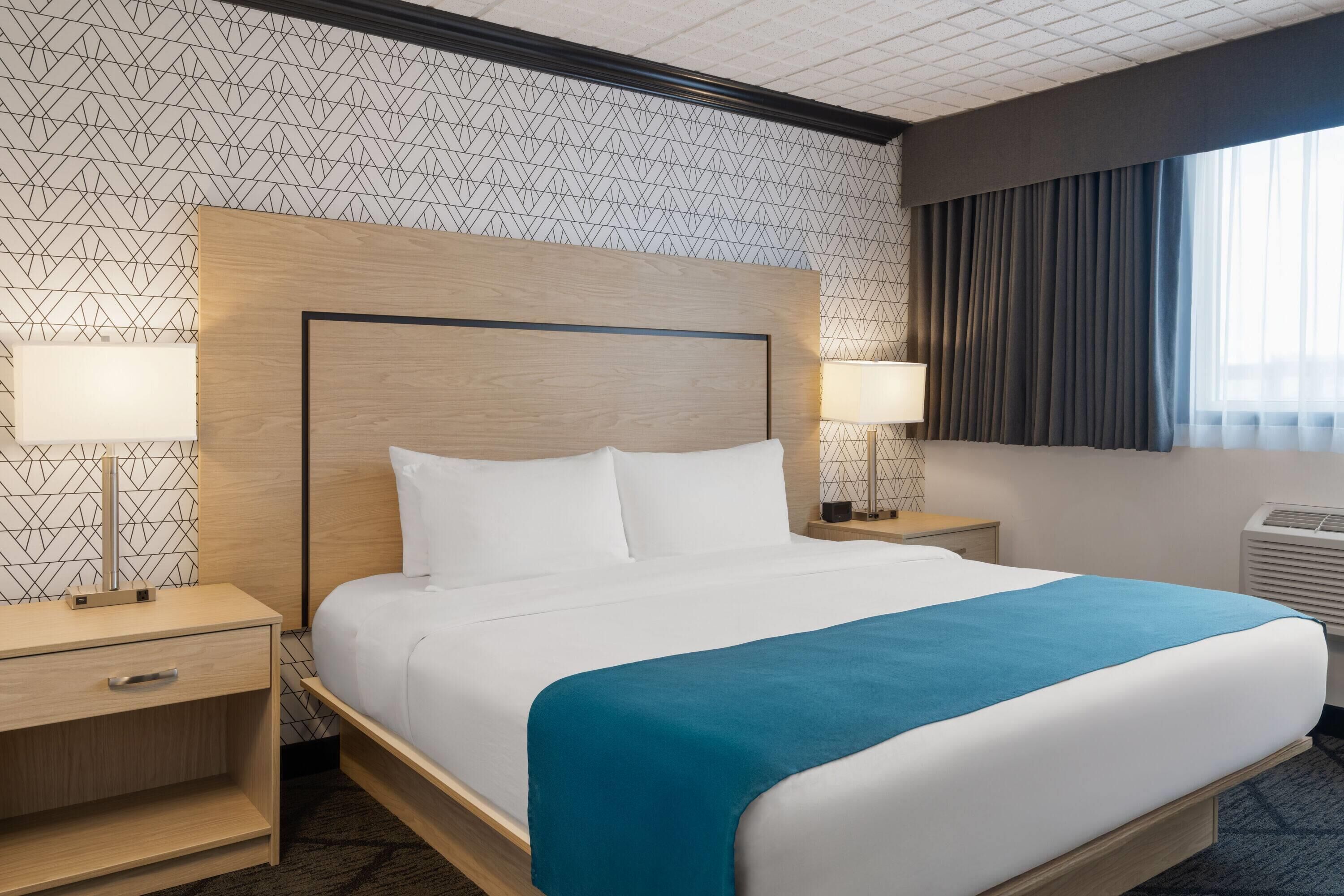 Une chambre d’hôtel avec un grand lit, deux tables de chevet avec des lampes, une tête de lit et une fenêtre avec des rideaux.
