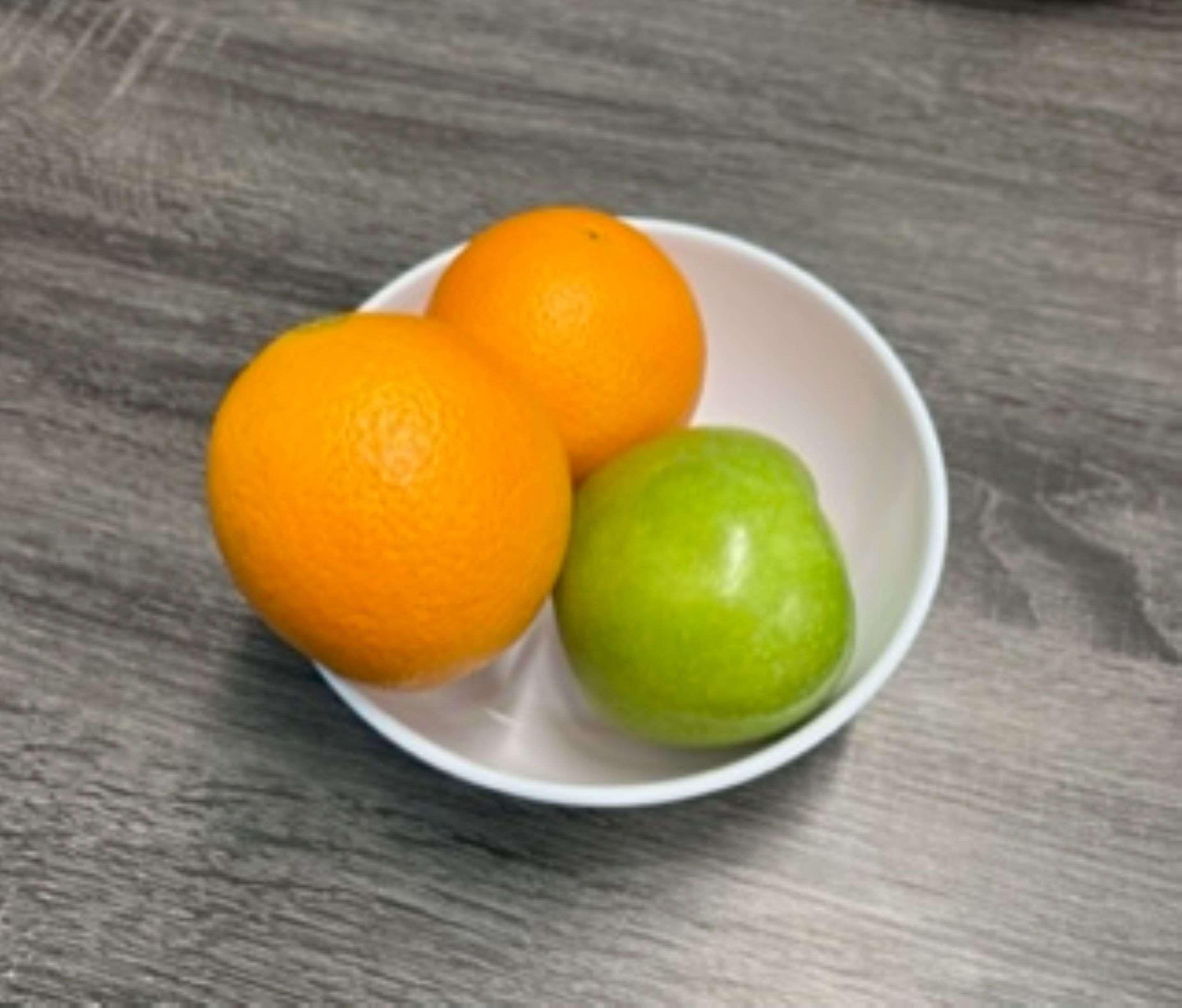 A bowl containing two oranges and one apple on a wooden surface.