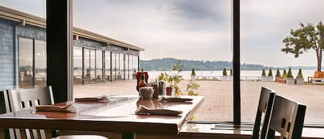 A dining area with a table set for two, overlooking a beach with a view of the water and distant hills.