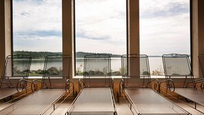 Une rangée de chaises en métal, dotées d’un dossier en treillis, est placée face à une grande fenêtre donnant sur un lac et une forêt.