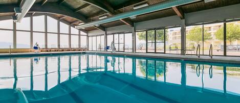 An indoor swimming pool with clear blue water, large windows, and a view of the outdoors.