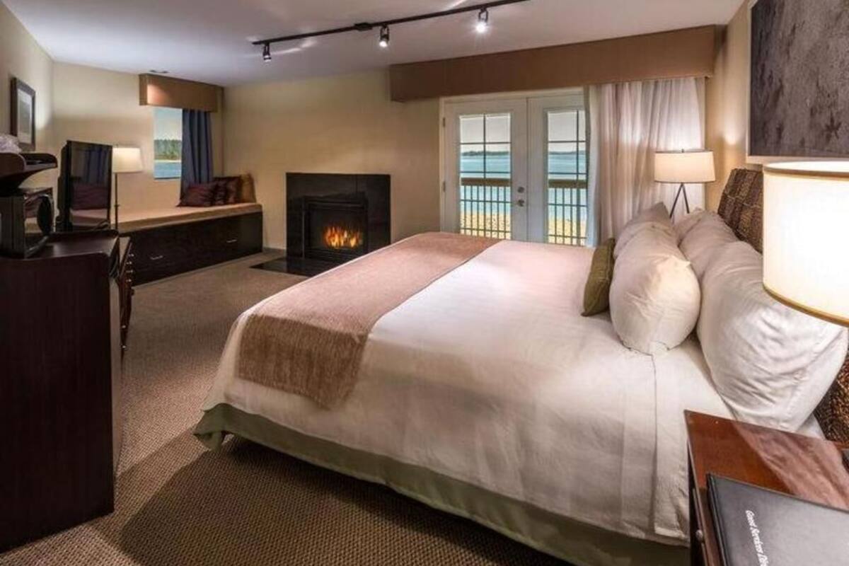 Een hotelkamer met een groot bed, een open haard, een bureau en uitzicht op het strand door de ramen.