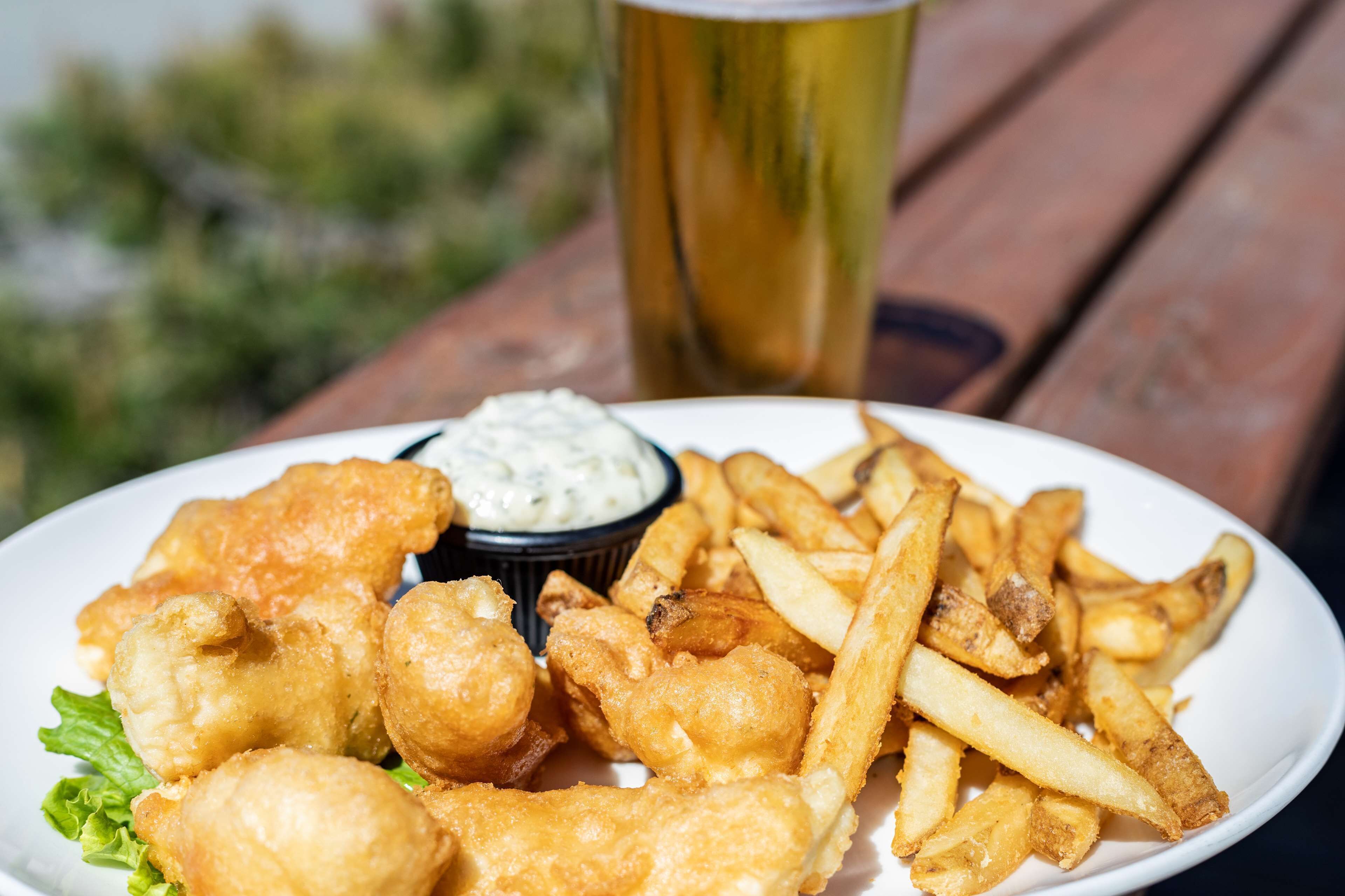 Un plato de pescado con papas fritas, salsa tártara y una cerveza en una mesa de madera.