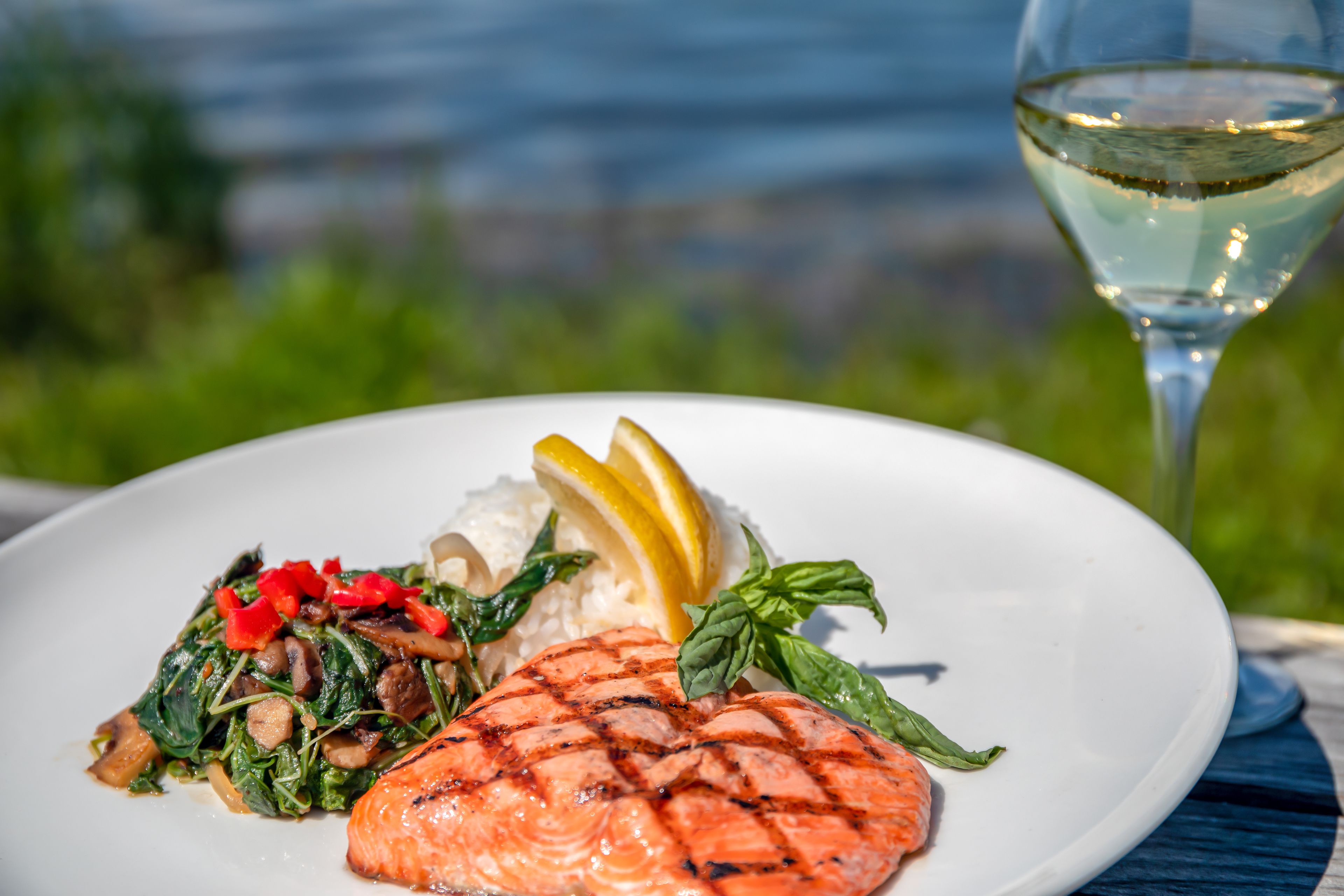 Un plato de comida con salmón a la parrilla, arroz y verduras, acompañado de una copa de vino blanco, sobre una mesa al aire libre con vistas.