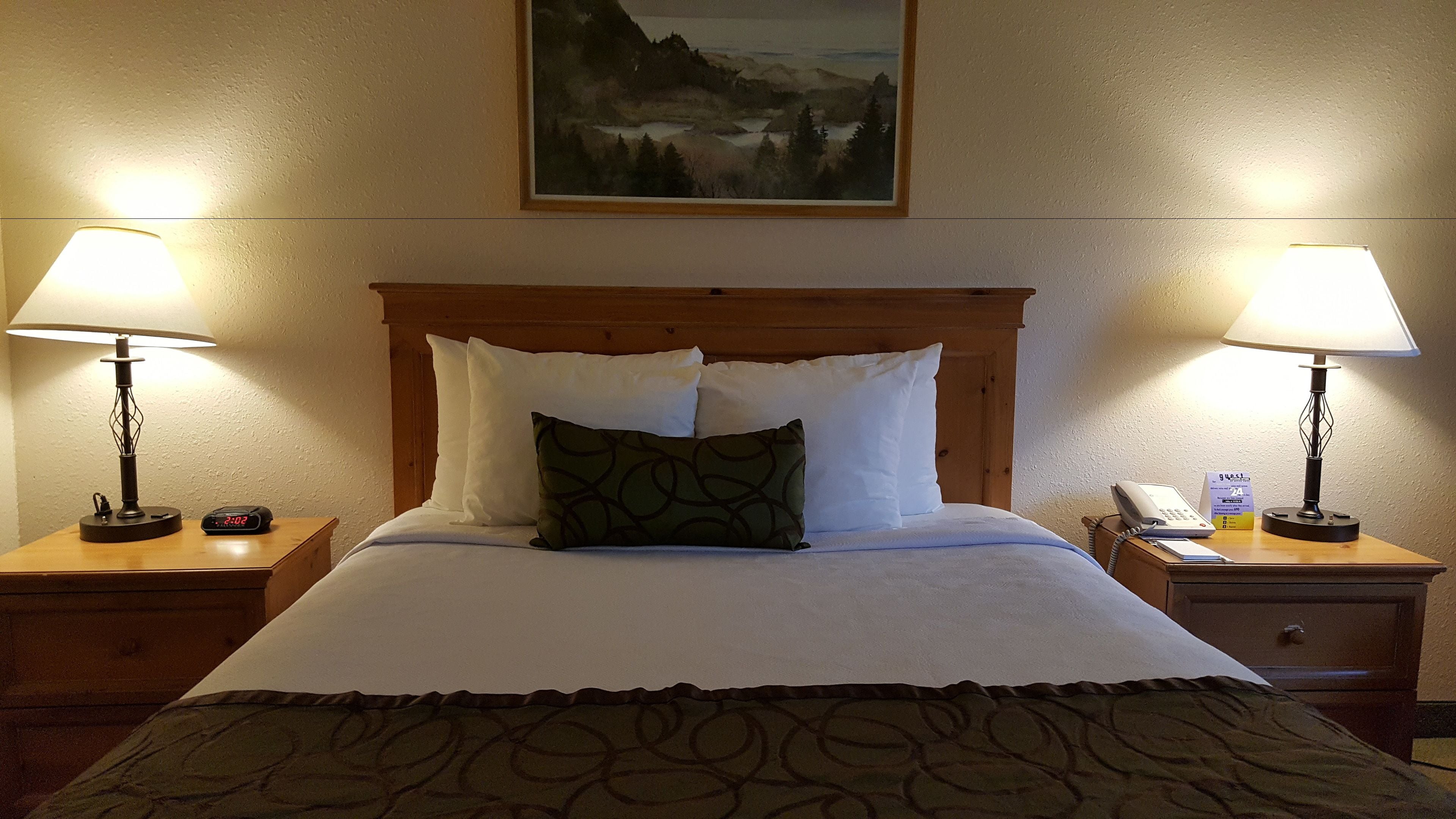 Une chambre d’hôtel avec un grand lit, deux lampes de chevet, un tableau au mur, une table de chevet avec un téléphone et une horloge, et une tête de lit en bois.