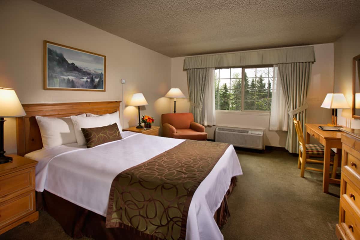 A hotel room with a bed, two bedside tables, a desk, a chair, a window with curtains, and a painting on the wall.