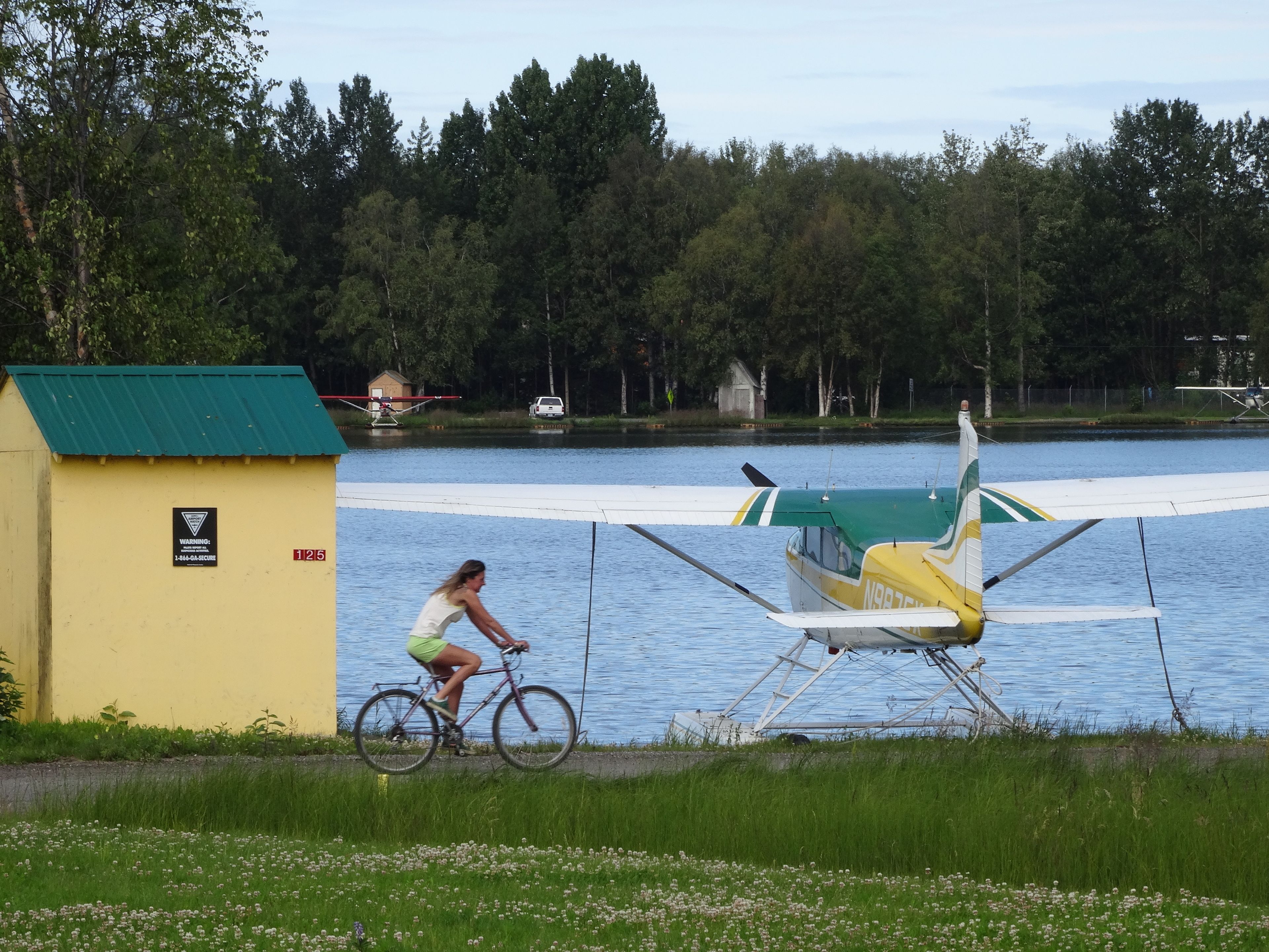 A woman riding a bicycle near a small yellow building with a green roof, a white and green airplane, and a lake with trees in the background.