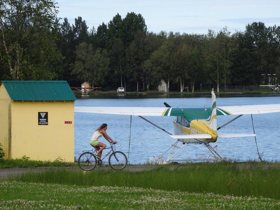 A woman riding a bicycle near a small yellow building with a green roof, a white and green airplane, and a lake with trees in the background.