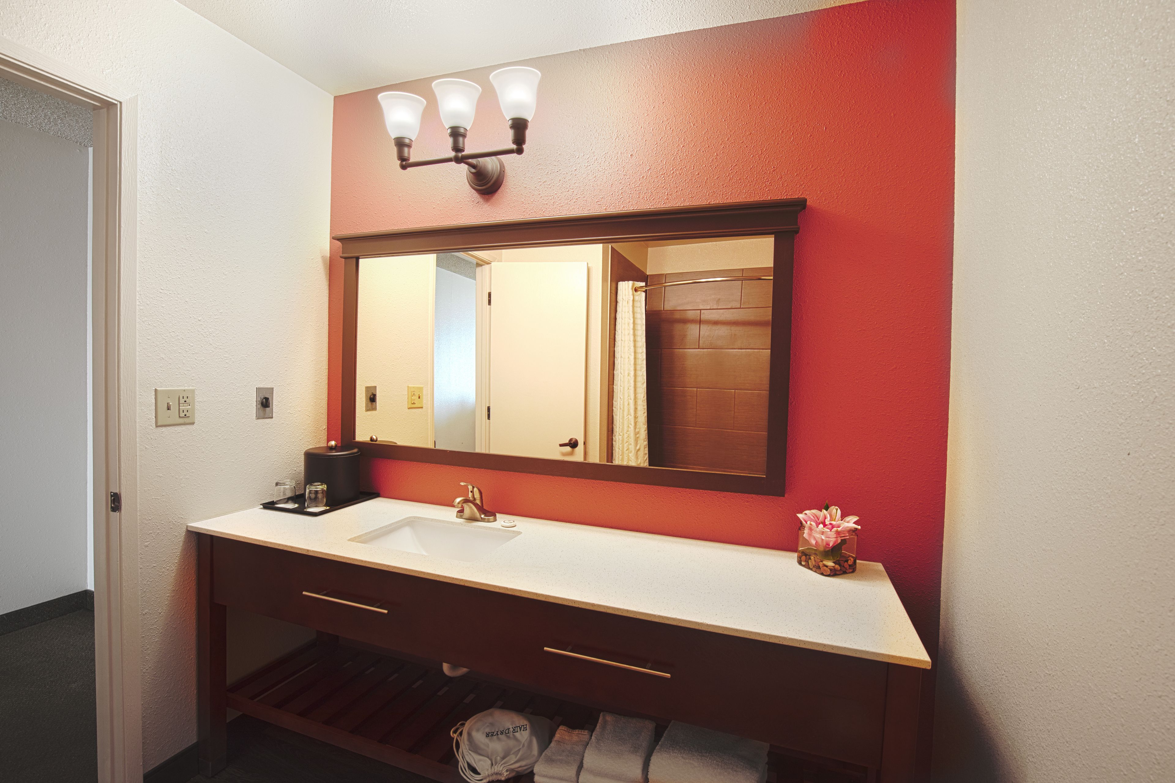 Une salle de bain avec un lavabo blanc, un miroir avec trois lampes et un mur rouge.