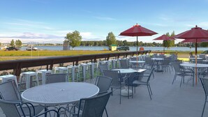 An outdoor patio with round tables and chairs, red umbrellas, and a view of a lake and mountains.