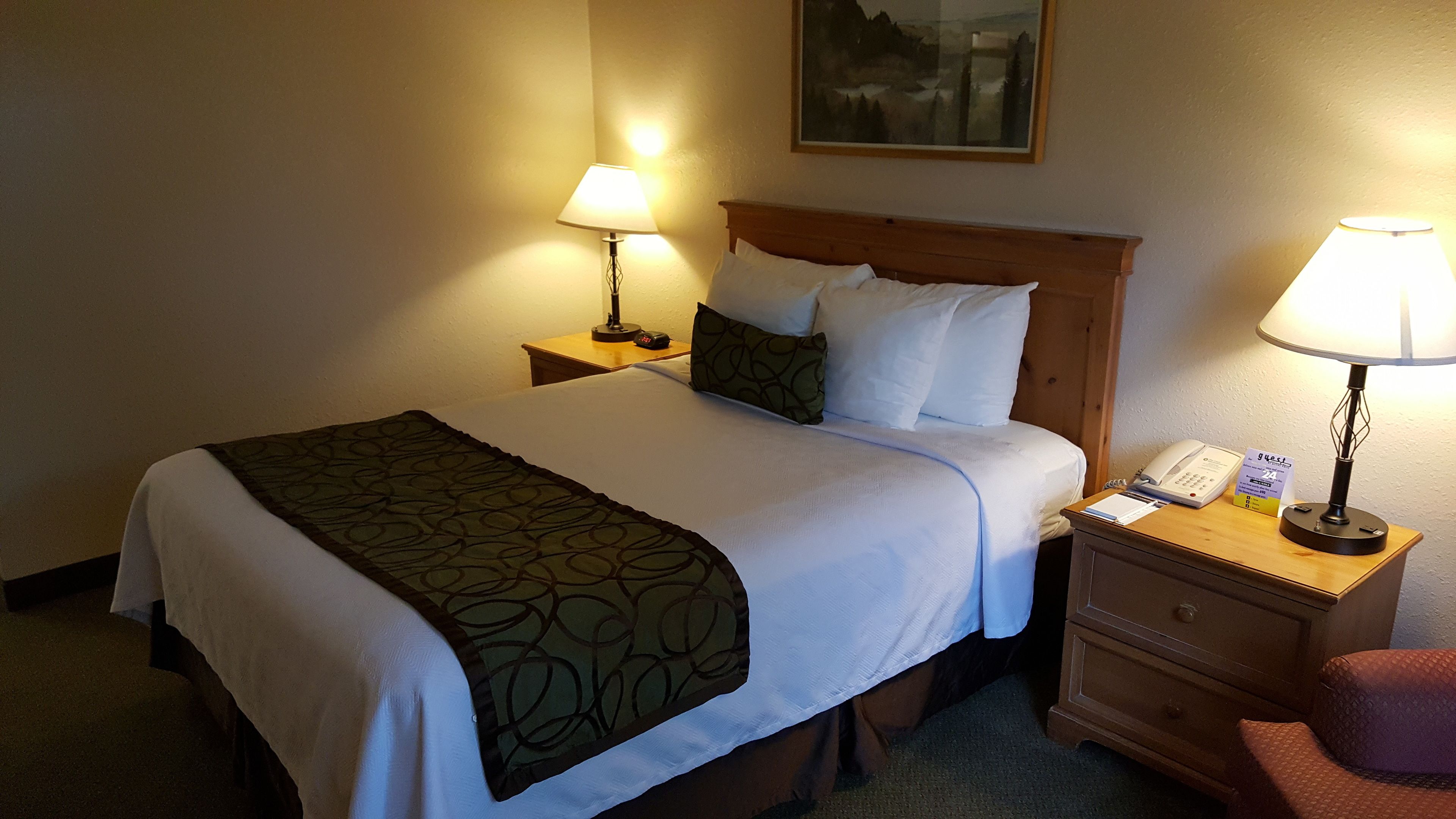 A hotel room with a neatly made bed, two bedside lamps, a wooden nightstand with a telephone and a notepad, and a chair.