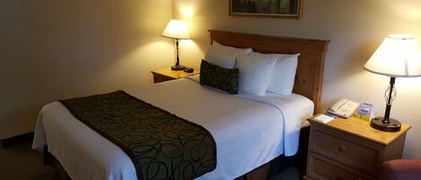 Habitación de hotel con una cama bien hecha, dos lámparas de noche, una mesita de madera con un teléfono y un bloc de notas, y una silla.
