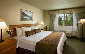 A hotel room with a large bed, two bedside tables with lamps, a window with curtains, and a painting on the wall.