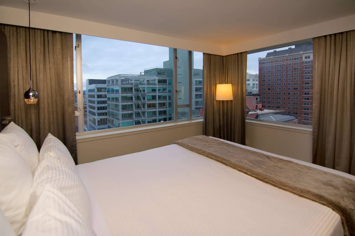 A hotel room with a large bed, a view of city buildings, and a lamp on the nightstand.