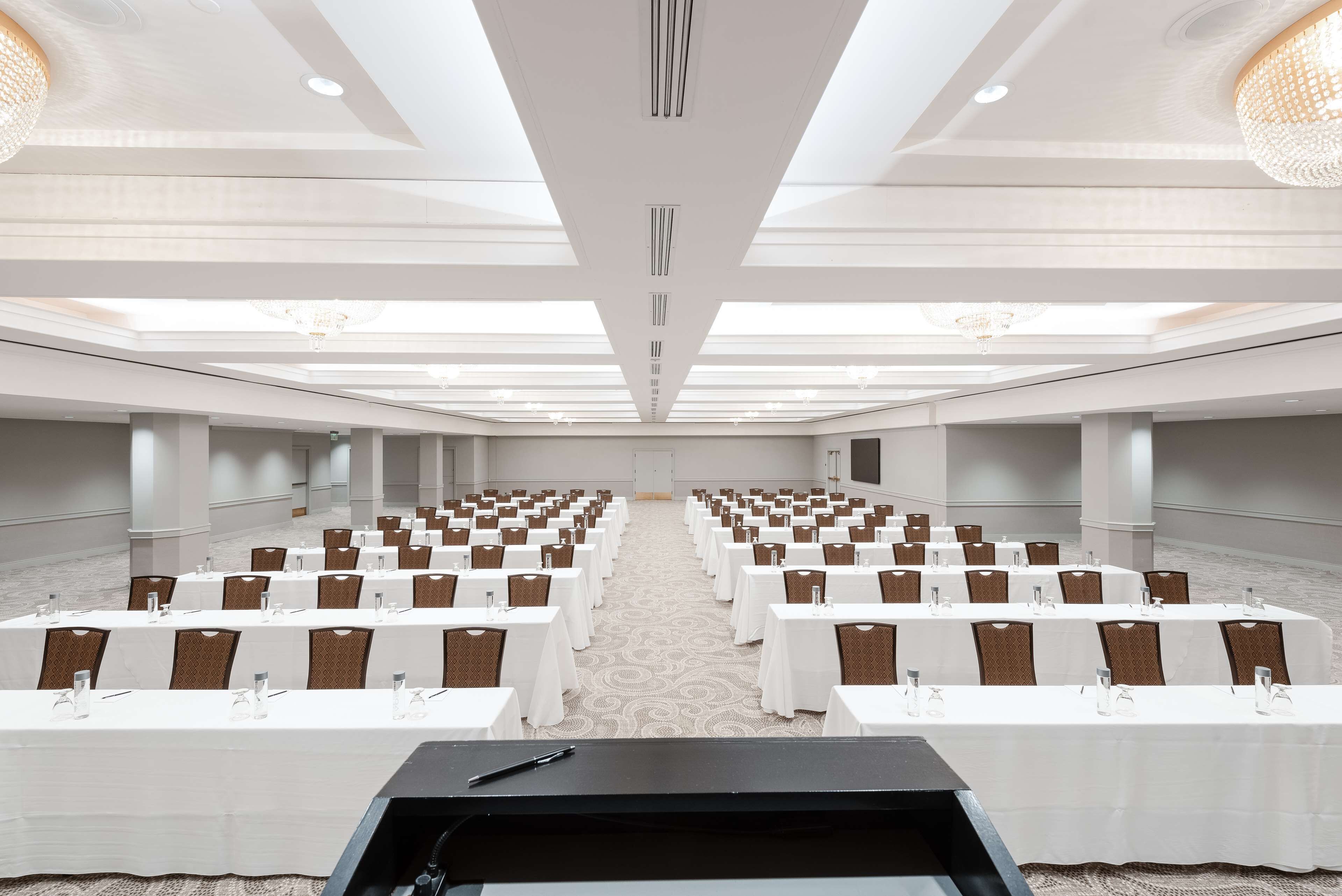 Un salón de conferencias con filas de mesas y sillas, un atril y suelo alfombrado.