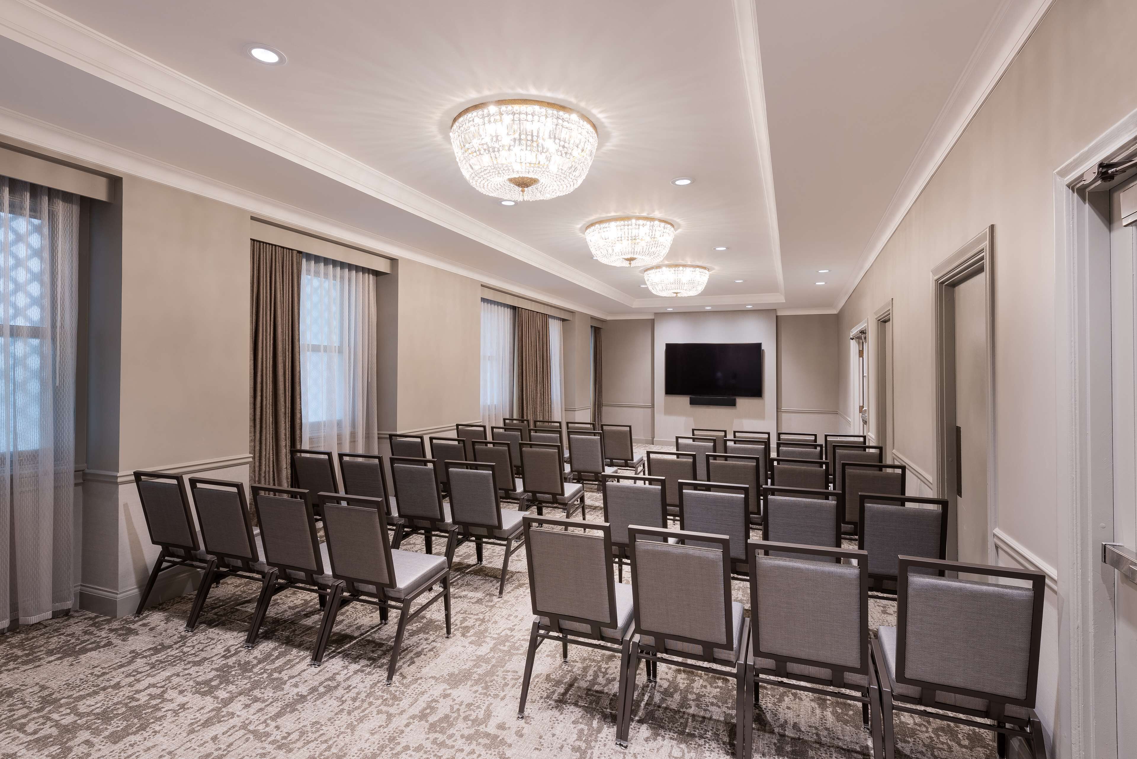 A conference room with rows of chairs, a chandelier, a flat-screen TV, and a carpeted floor.