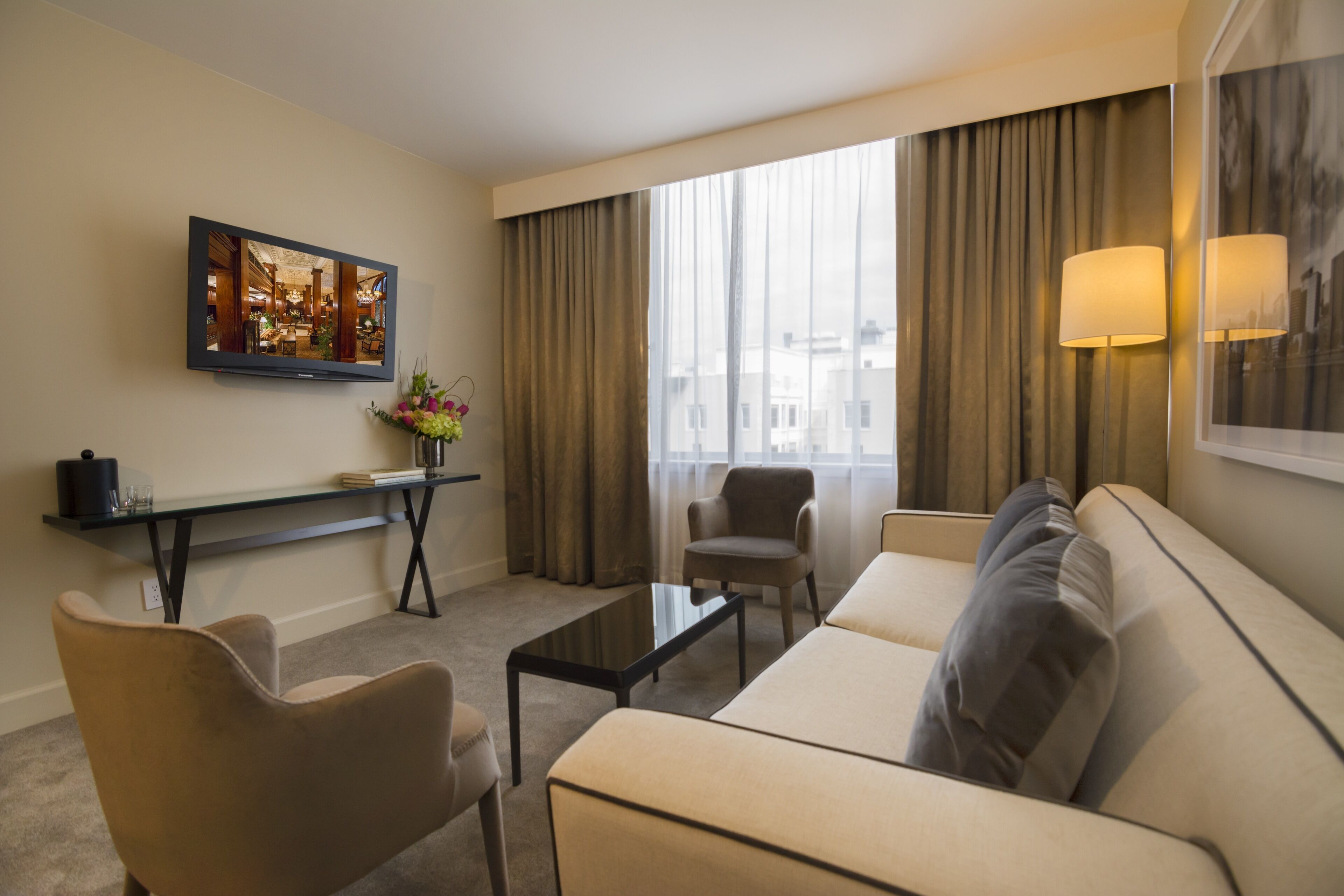 Una habitación de hotel moderna con sofá, sillones, una mesita de centro, televisión y un gran ventanal con cortinas.