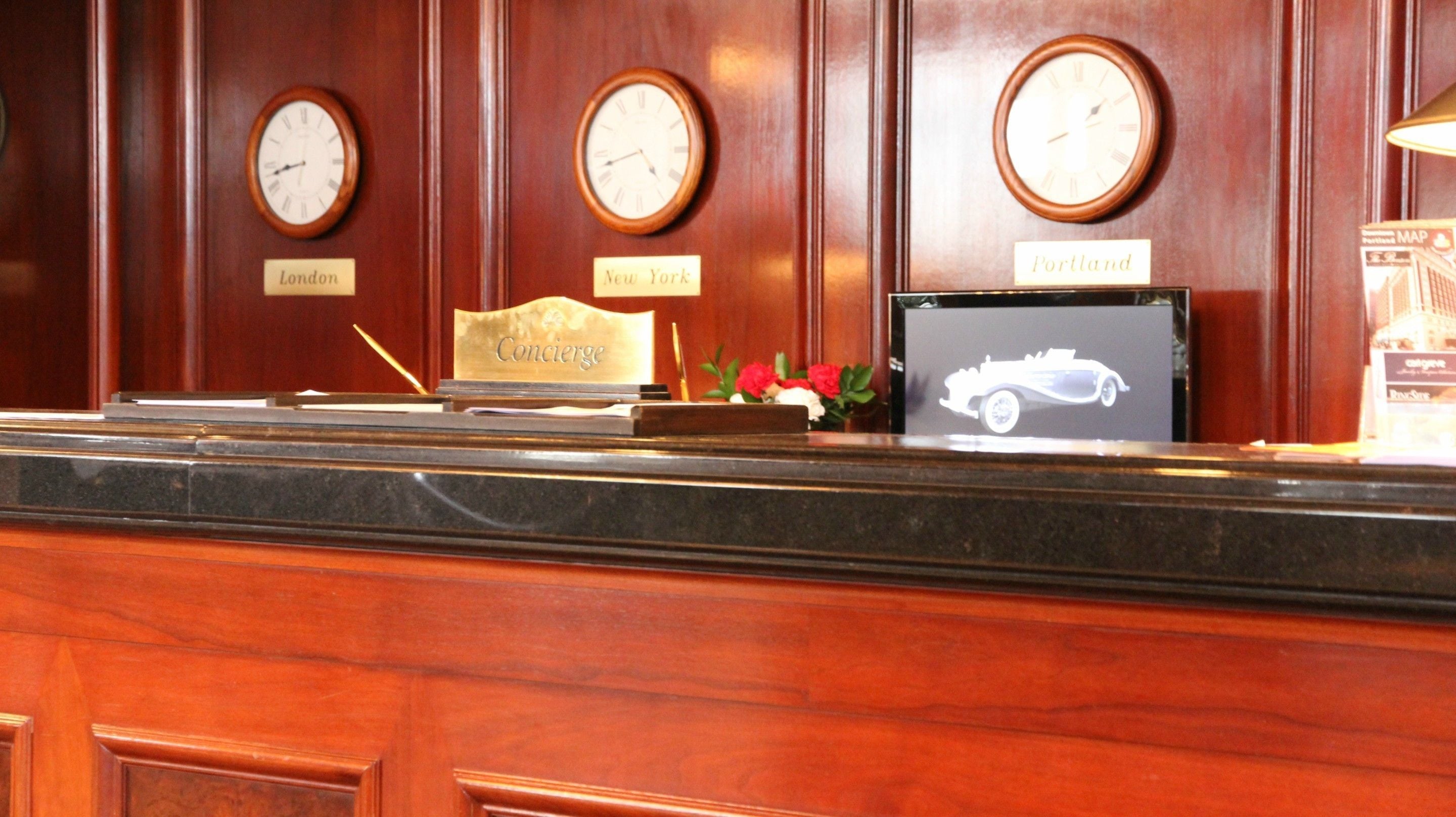 A reception desk with clocks labeled London, New York, and Portland, a framed picture of a car, and a small bouquet of flowers.