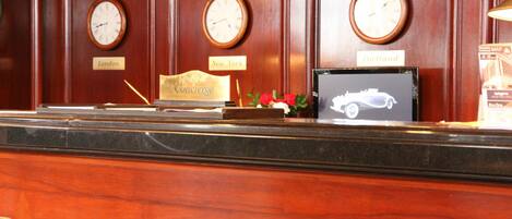 A reception desk with clocks labeled London, New York, and Portland, a framed picture of a car, and a small bouquet of flowers.