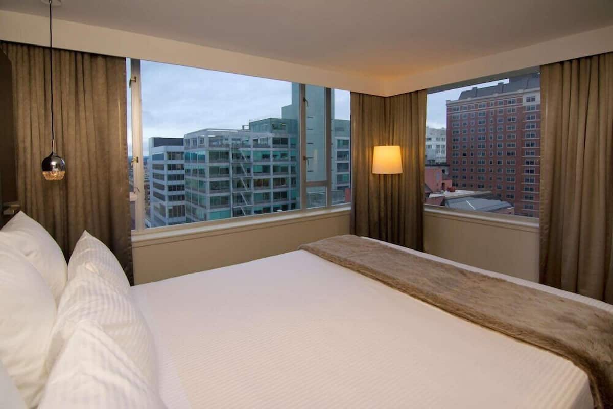 A hotel room with a large bed, a window with curtains, a lamp, and a view of buildings.