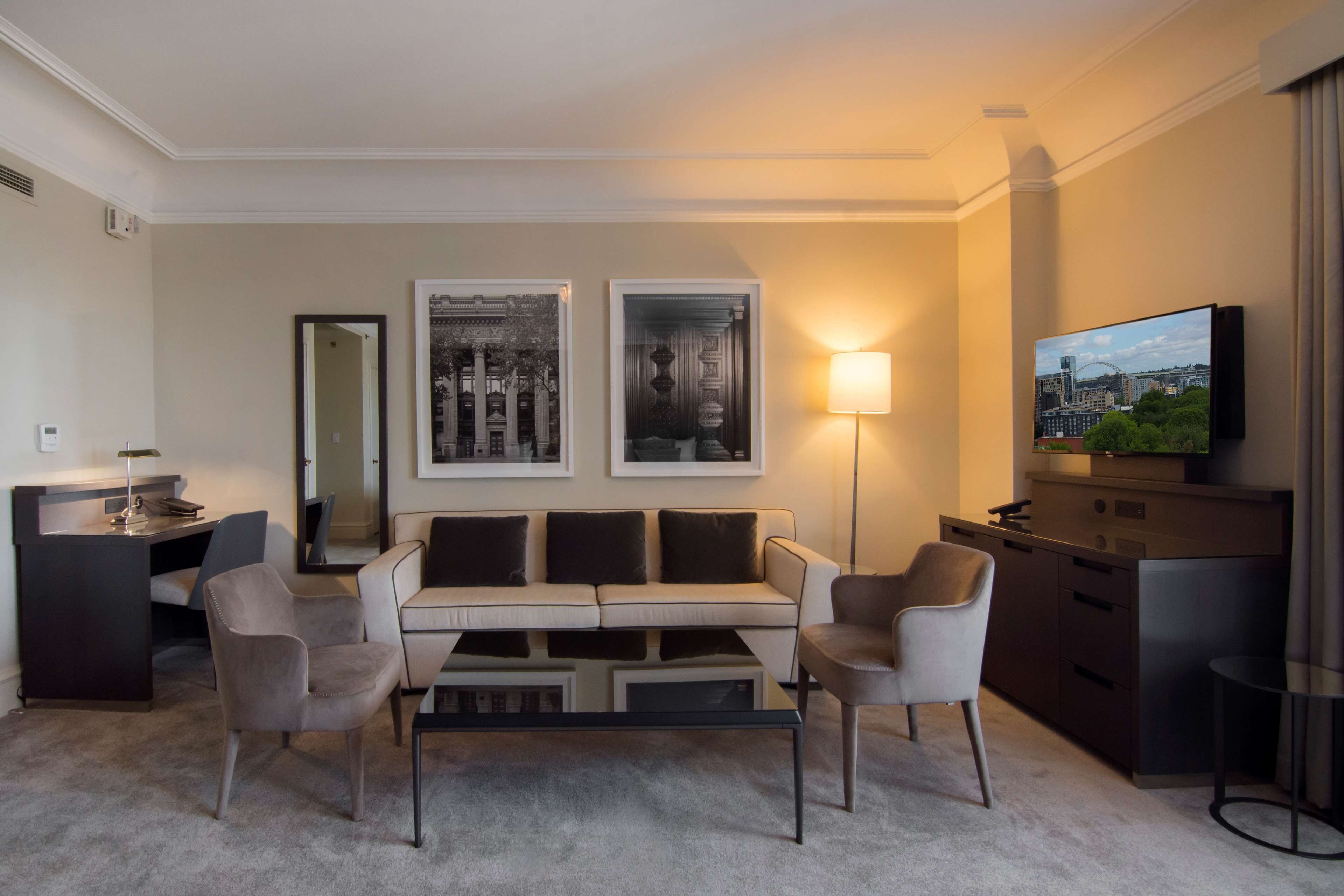 Et moderne hotelværelse med sofa, lænestole, sofabord, skrivebord og fjernsyn.