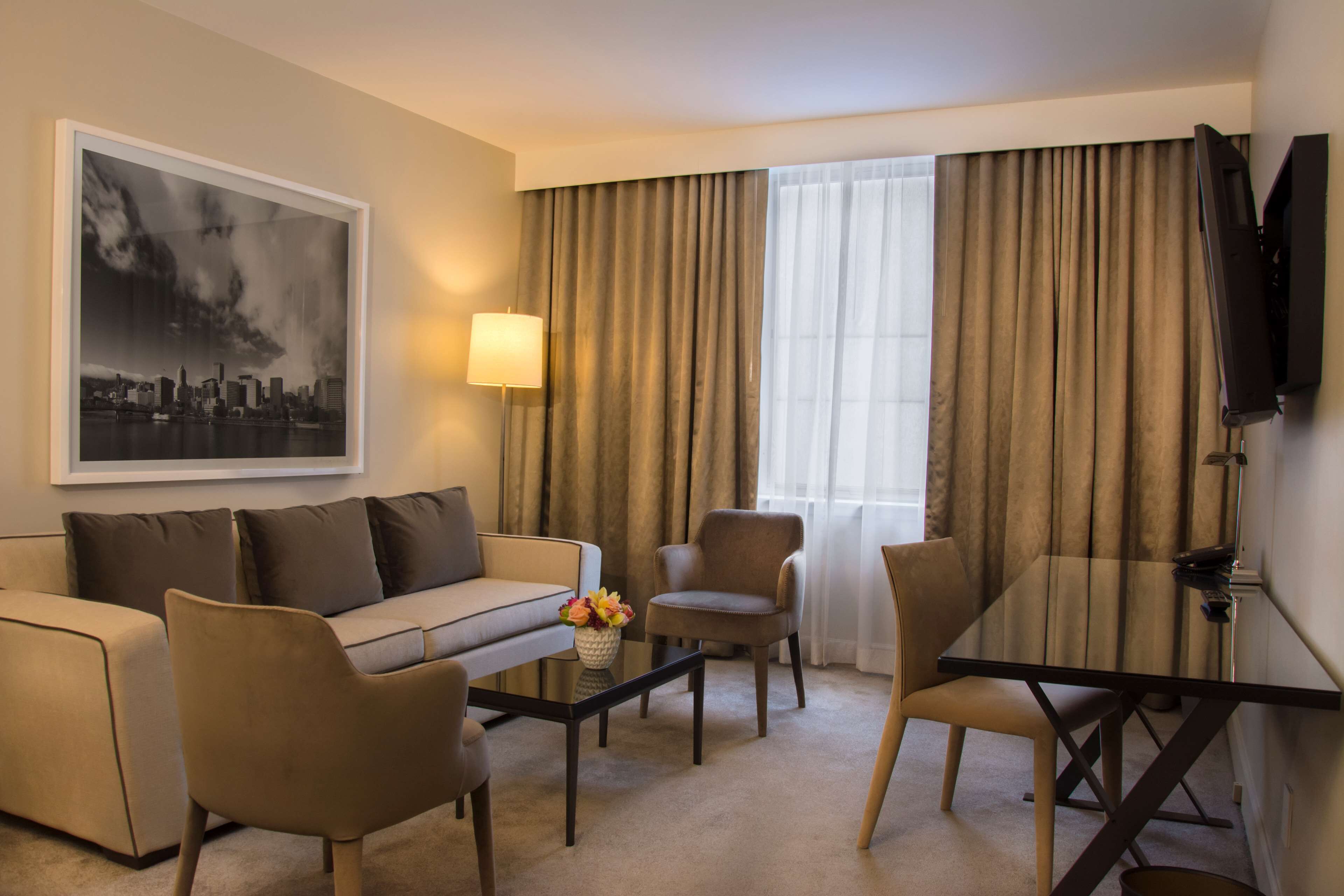 Une chambre d’hôtel avec un canapé, des fauteuils, une table basse ornée de fleurs, un téléviseur et des rideaux.