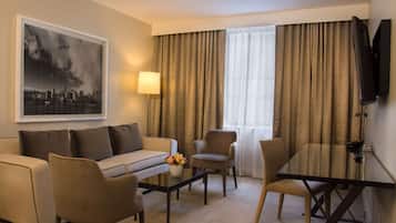 Une chambre d’hôtel avec un canapé, des fauteuils, une table basse ornée de fleurs, un téléviseur et des rideaux.