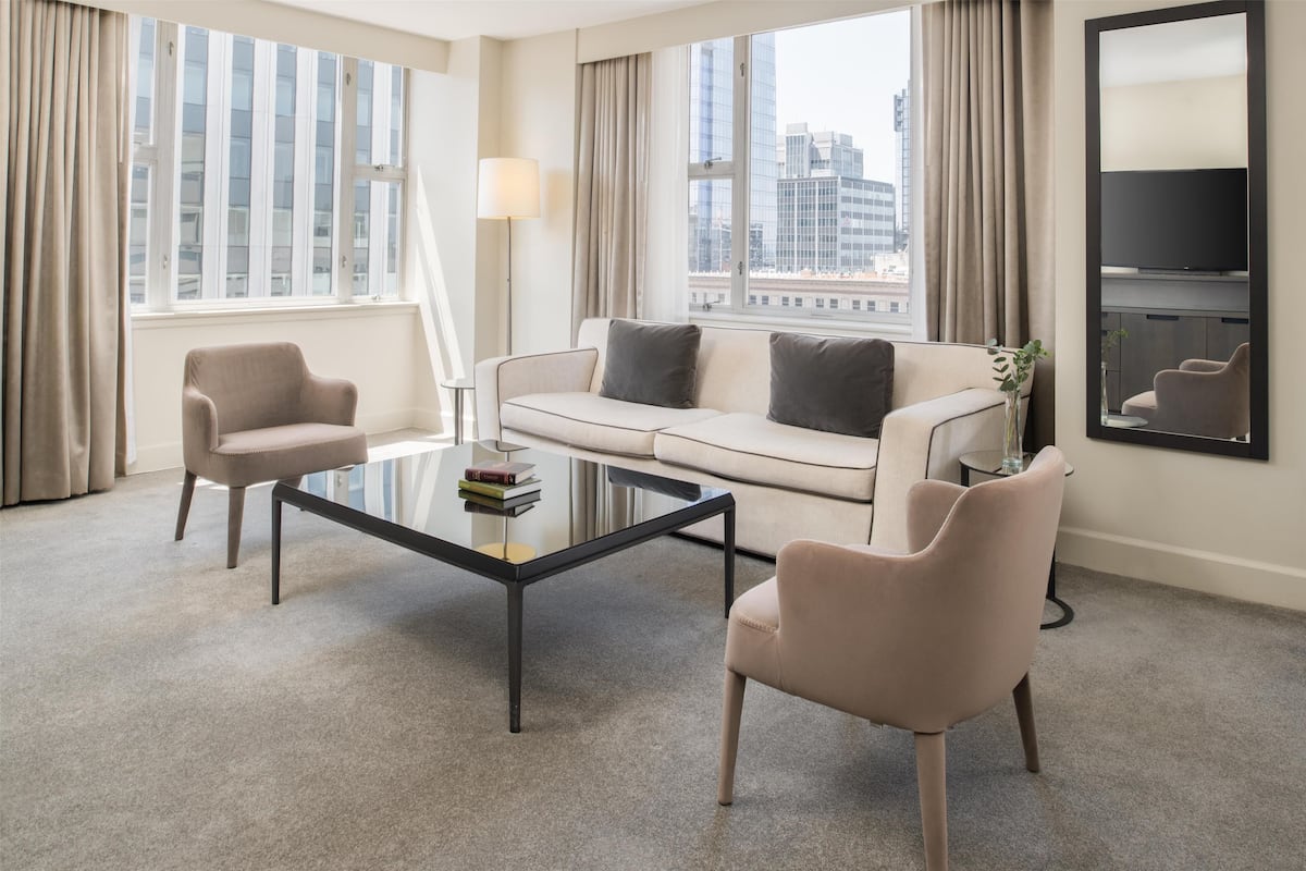A modern hotel room with a sofa, two armchairs, a glass coffee table, and a large window offering a city view.