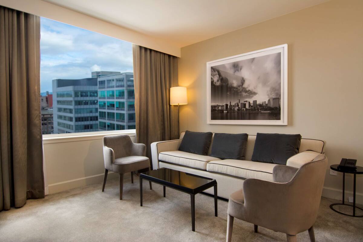 A hotel room with a sofa, two armchairs, a coffee table, a lamp, a framed cityscape picture, and a window with curtains.
