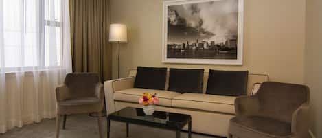 Una habitación de hotel con un sofá color beige, dos sillones, una mesa de centro con un jarrón de flores, una lámpara de pie y un cuadro de un paisaje urbano en la pared.