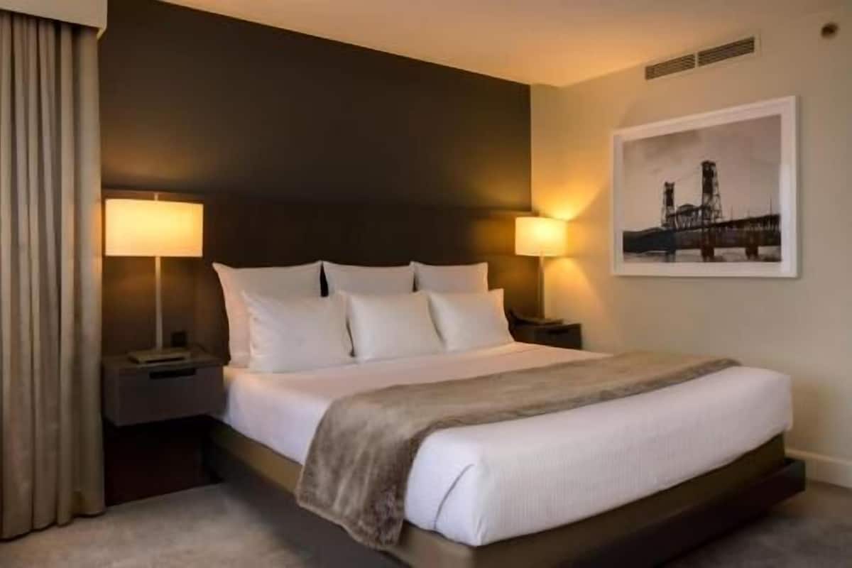 A hotel room with a large bed, two bedside lamps, a headboard, a framed picture on the wall, and a window with curtains.