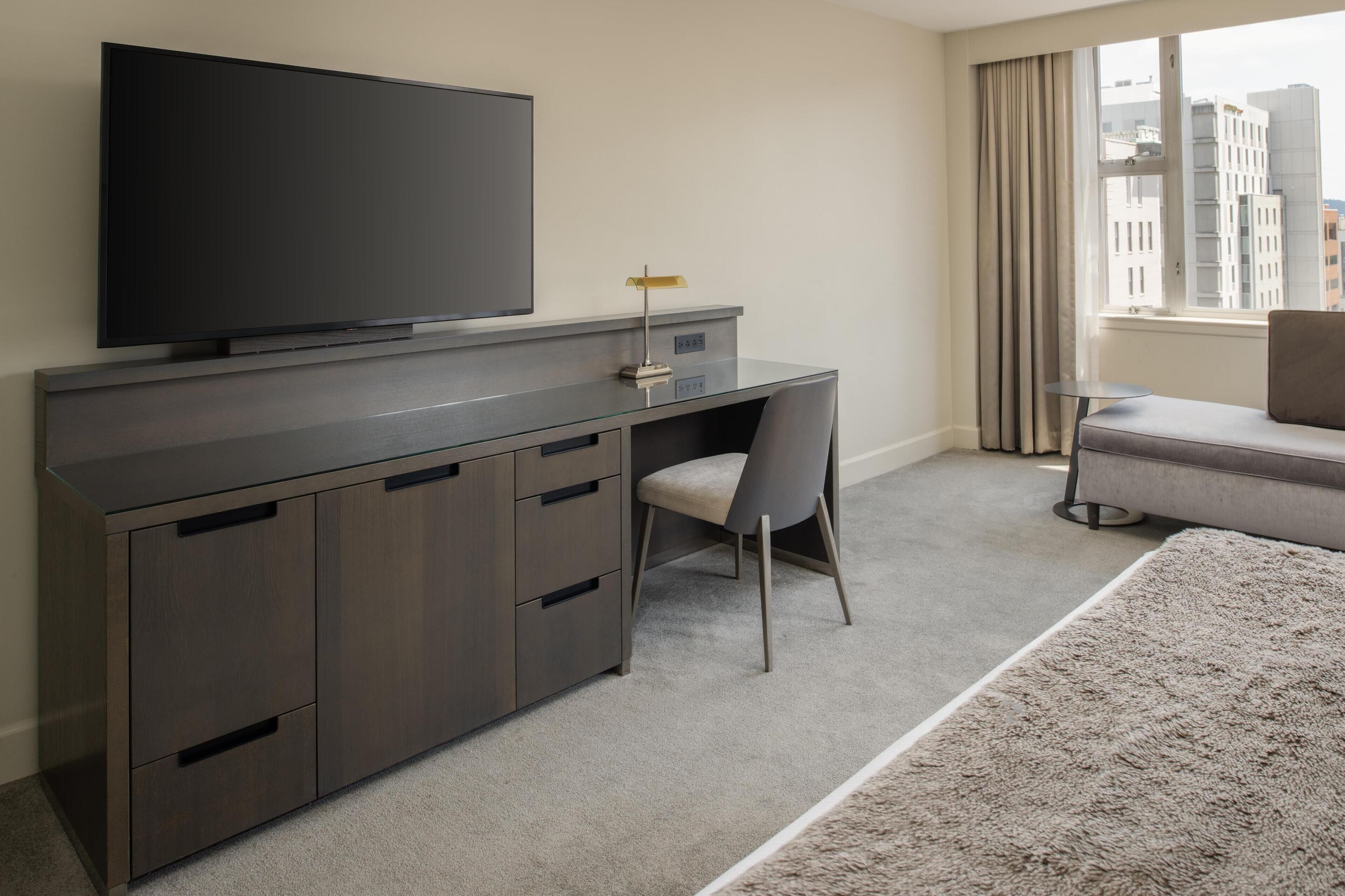 Une chambre d’hôtel moderne, dotée d’un grand téléviseur à écran plat fixé sur un meuble en bois foncé, d’un fauteuil de bureau gris, d’un petit bureau avec une lampe, d’un canapé gris et offrant une vue sur les bâtiments à travers une fenêtre ornée de rideaux beige.