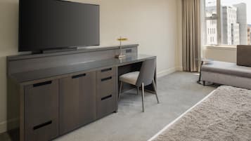 Une chambre d’hôtel moderne, dotée d’un grand téléviseur à écran plat fixé sur un meuble en bois foncé, d’un fauteuil de bureau gris, d’un petit bureau avec une lampe, d’un canapé gris et offrant une vue sur les bâtiments à travers une fenêtre ornée de rideaux beige.