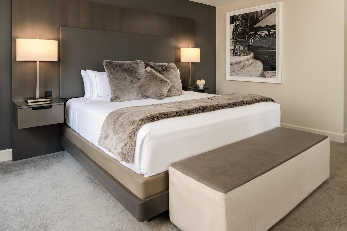 A modern bedroom with a large bed, two bedside lamps, a nightstand with a book, a bench, and a framed picture on the wall.