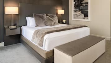 A modern bedroom with a large bed, two bedside lamps, a nightstand with a book, a bench, and a framed picture on the wall.