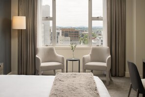 A hotel room with a large bed, two armchairs, a small table with a vase, and a view of a cityscape through large windows.