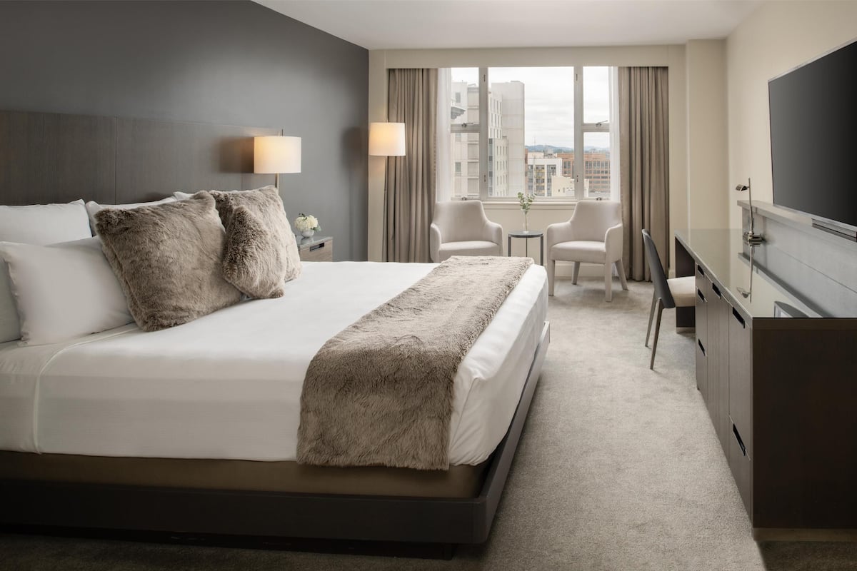A hotel room with a large bed, two bedside lamps, a desk with a chair, and a view of the city through large windows.