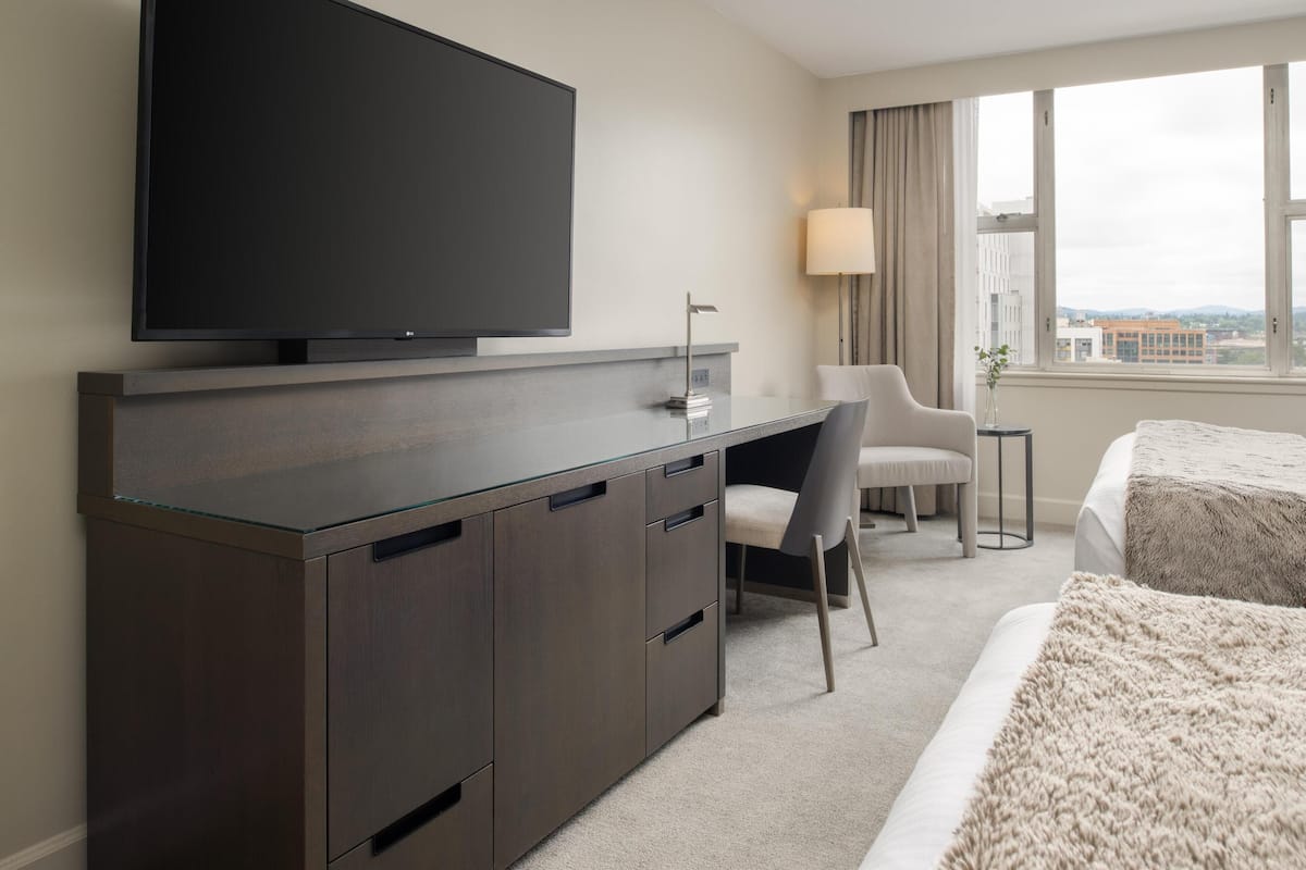 A modern hotel room with a flat-screen TV mounted on a dark wooden desk, a comfortable chair, a bedside table with a lamp, and a large window offering a city view.