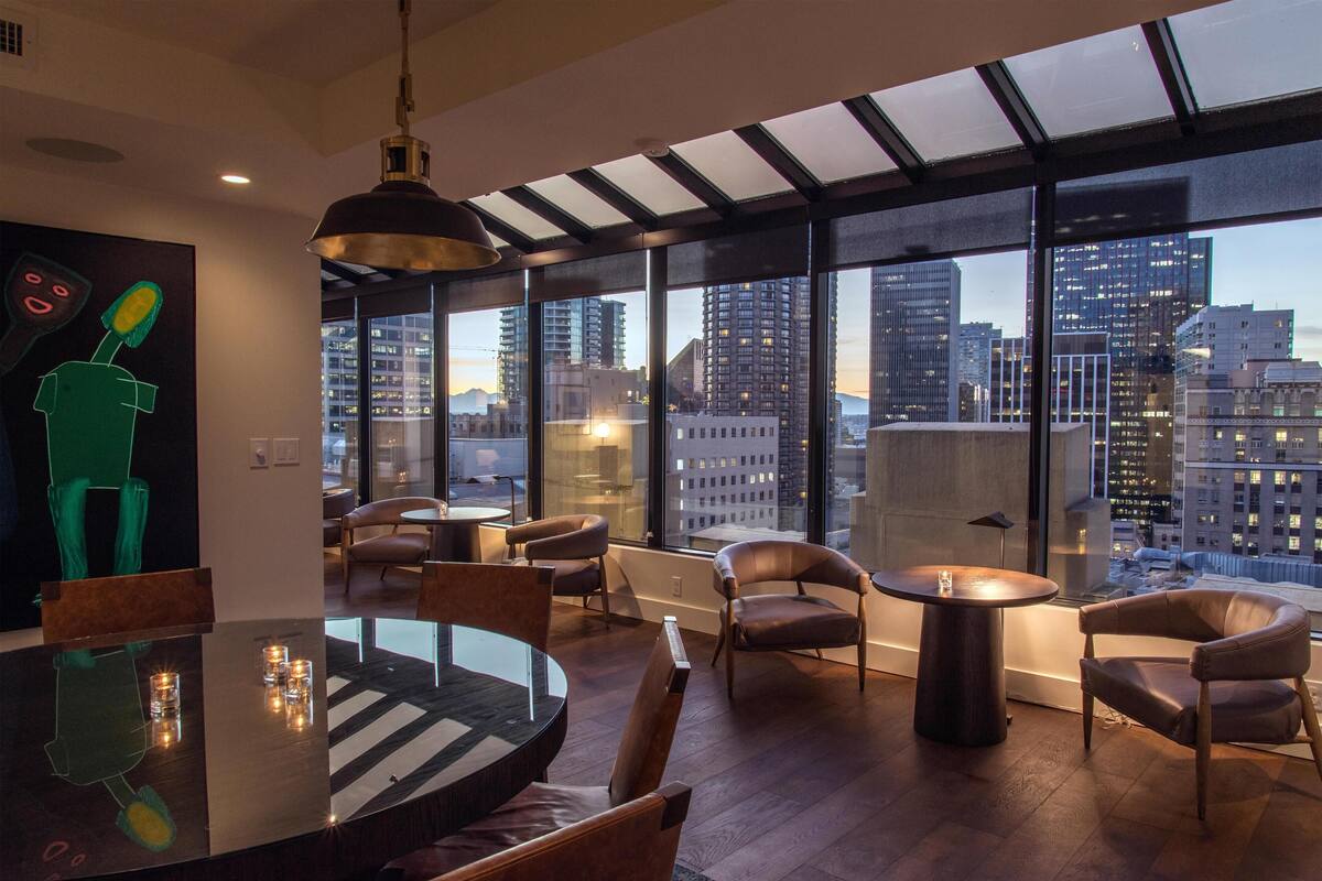 A modern dining area with a large table and chairs, large windows with a city view, and a unique green sculpture on the wall.