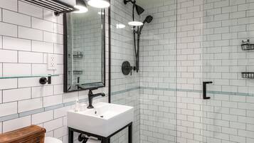 A modern bathroom with a white tiled wall, a glass shower enclosure, a white sink, and a wooden vanity.