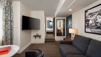A hotel room with a flat-screen TV, a sofa, a dining table, and a bed in the background.