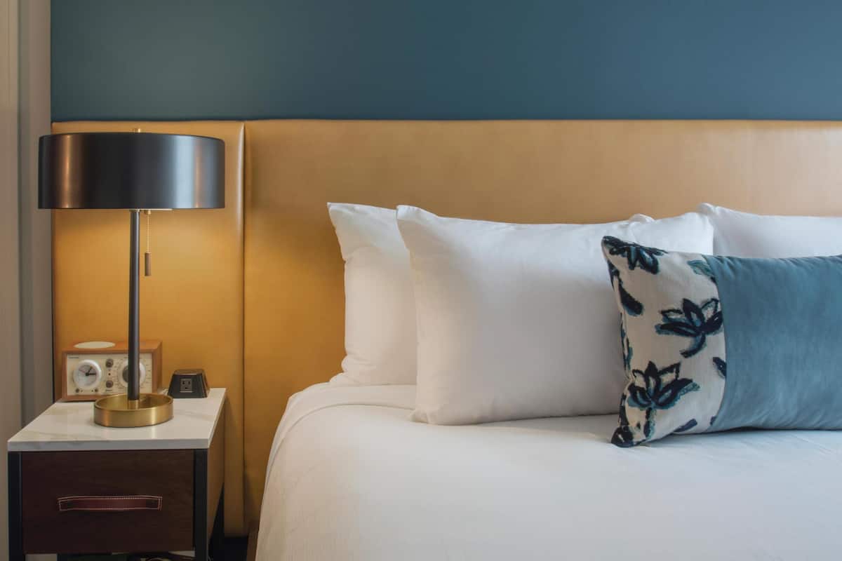 A neatly made bed with white and blue floral pillows, a bedside table with a lamp and a radio, and a wall with a teal and beige color scheme.