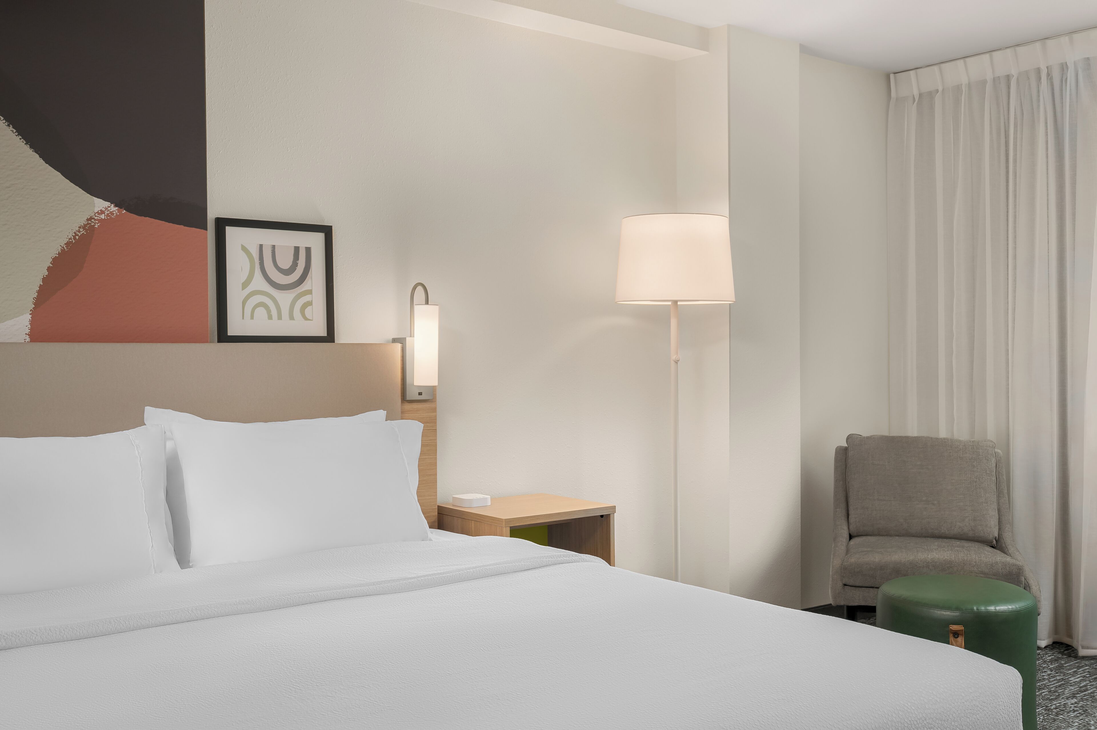 Habitació d'hotel amb un llit, una cadira, una làmpada, una taula de nit i una paret amb un quadre parcial.