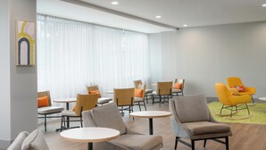 A modern hotel lobby with round tables and chairs, a large window with sheer curtains, and a yellow rocking chair. - Spark by Hilton Seattle Airport (SeaTac)