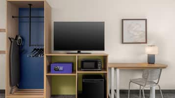 Una habitación con televisión, lámpara, silla, microondas, cubo de basura y perchero con ganchos.